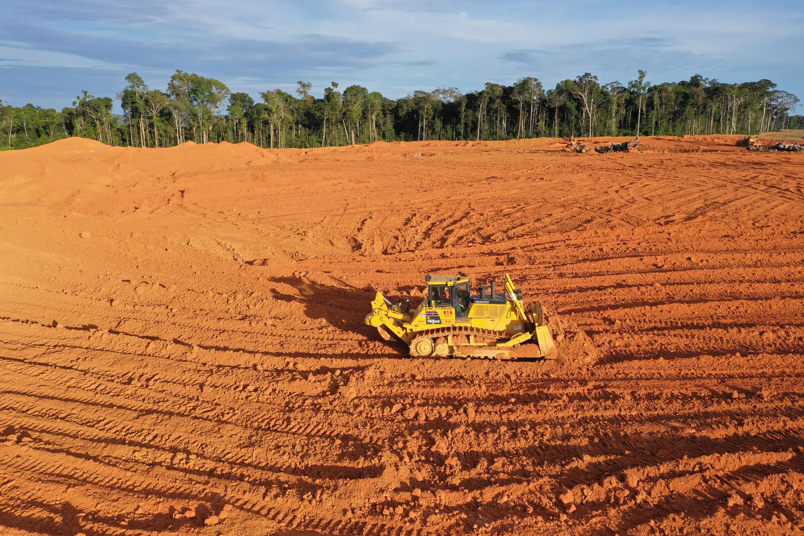 Trator de esteira espalhando terra vegetal em área aberta, com a floresta ao fundo
